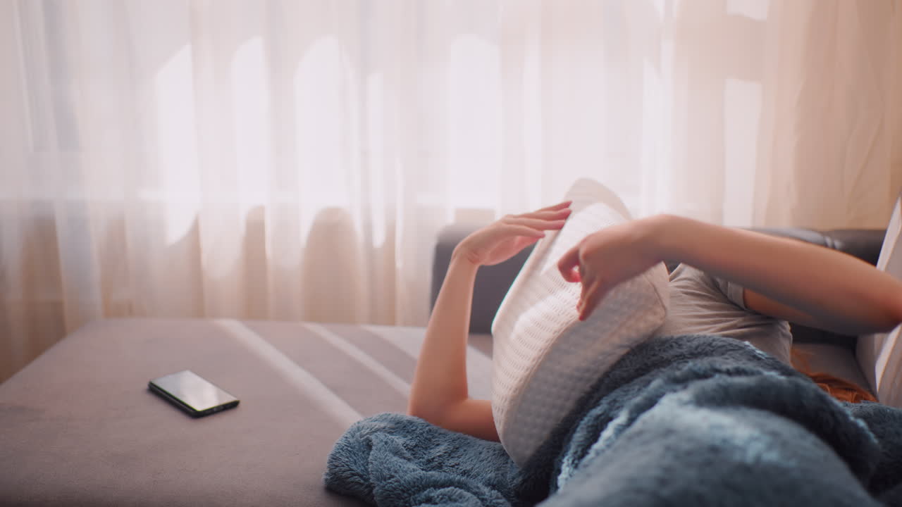Exhausted marketer rests on sofa pressing pillow against face under morning light with phone close by on couch, unwilling to get up after workload, seeking brief escape from stress