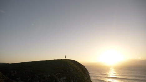 hombre caminando a lo largo del acantilado costero al amanecer con una tabla de surf