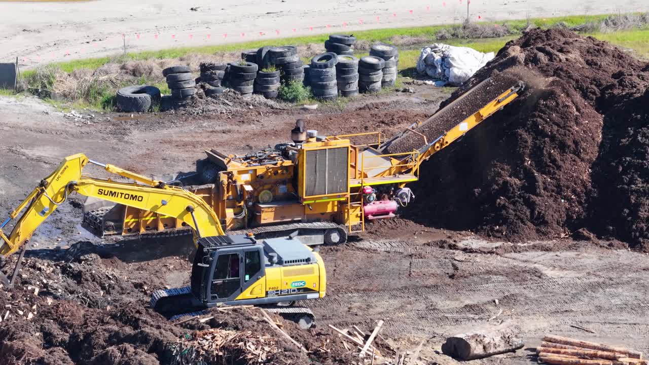 Aerial footage of an excavator loading mulch into a processing machine, surrounded by piles of soil and tires
