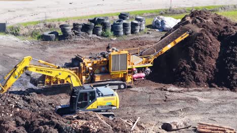 aerial footage of an excavator loading mulch into a processing machine, surrounded by piles of soil and tires