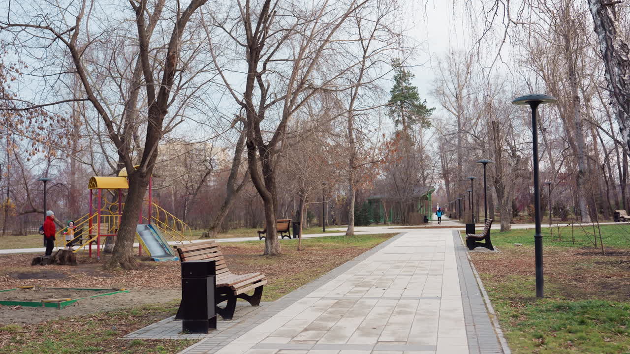 Recreational park view with sitting benches, children playing at playground area, and people walking along paved path surrounded by bare winter trees under cloudy sky in calm urban atmosphere