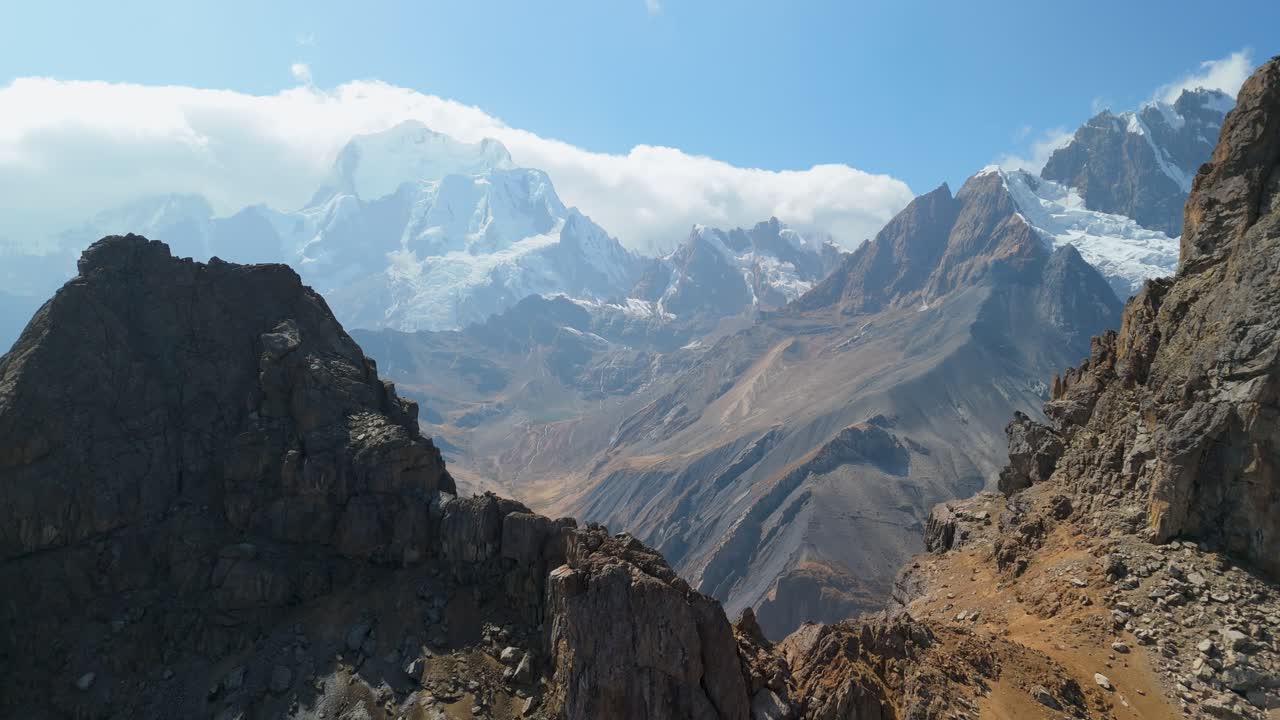 A breathtaking aerial pull-back shot reveals the immense scale of a remote mountain valley and the majestic, snow-capped peaks of the Huayhuash trek in the Peruvian Andes