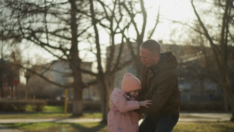 a man wearing a brown jacket carries a little girl in a pink cap and jacket as he gently lowers her to the ground in a park setting, surrounded by trees and a sunny background