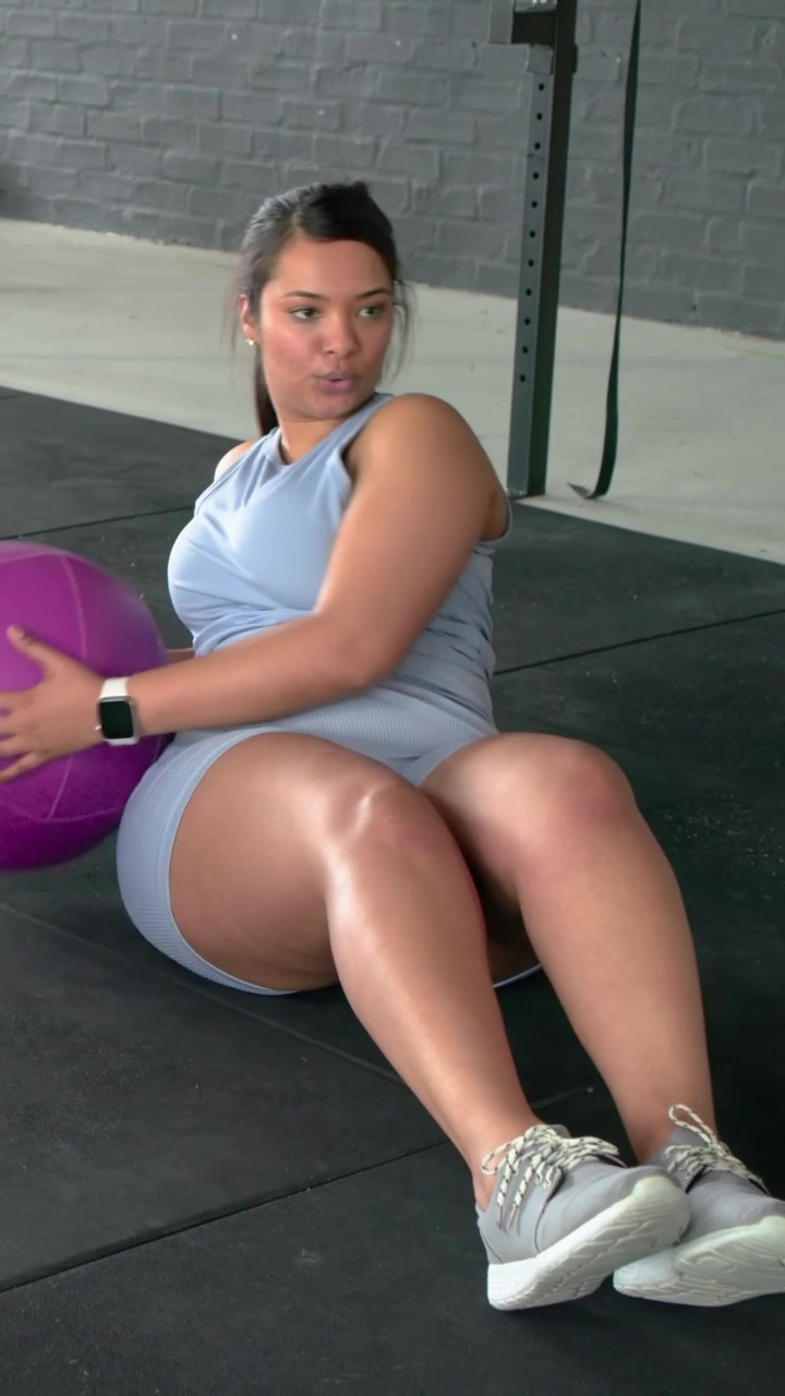 Video verticale: Donna con top blu che si torce, tiene una palla viola da 10 libbre per il core in palestra, strofinandosi la coscia.