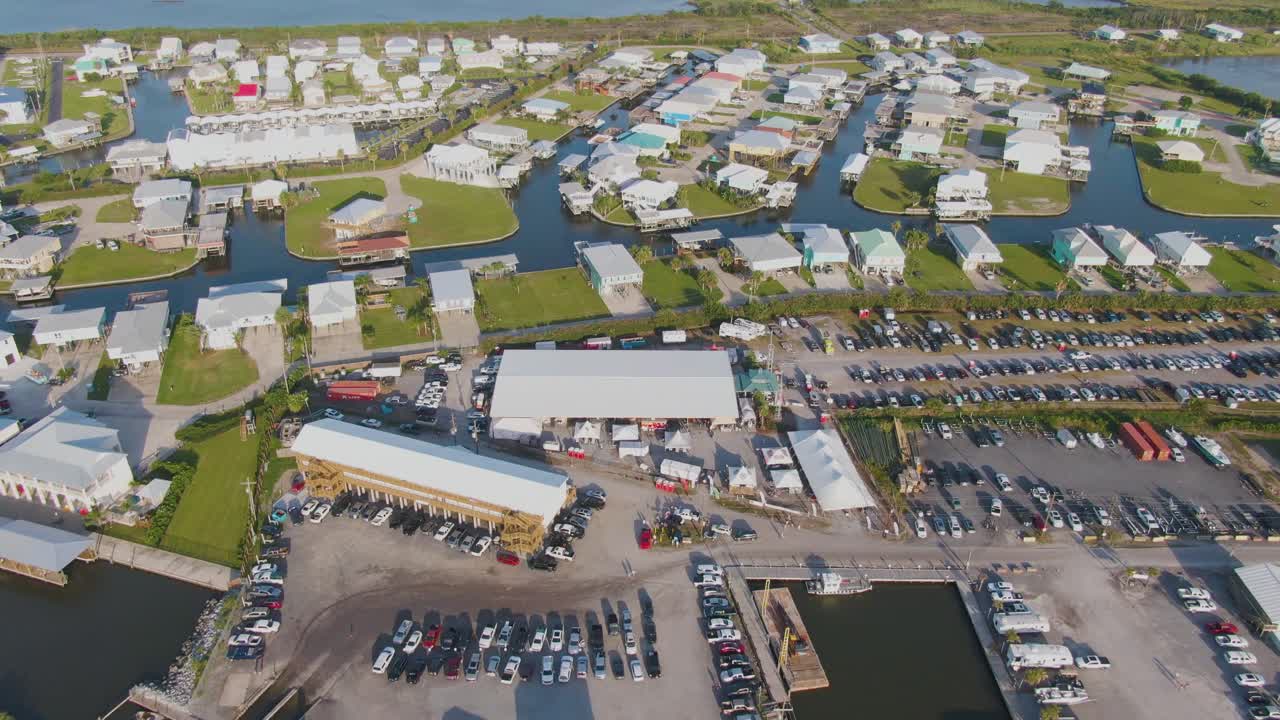 Aerial drone zooming out over Tarpon Rodeo Event Center in Grand Isle during music festival. Shows crowd, full parking lot, marina with boats, stilt homes, green grass, and nearby construction site