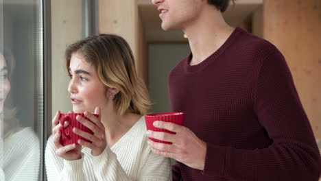 couple enjoying hot drinks by window, feeling cozy at home together