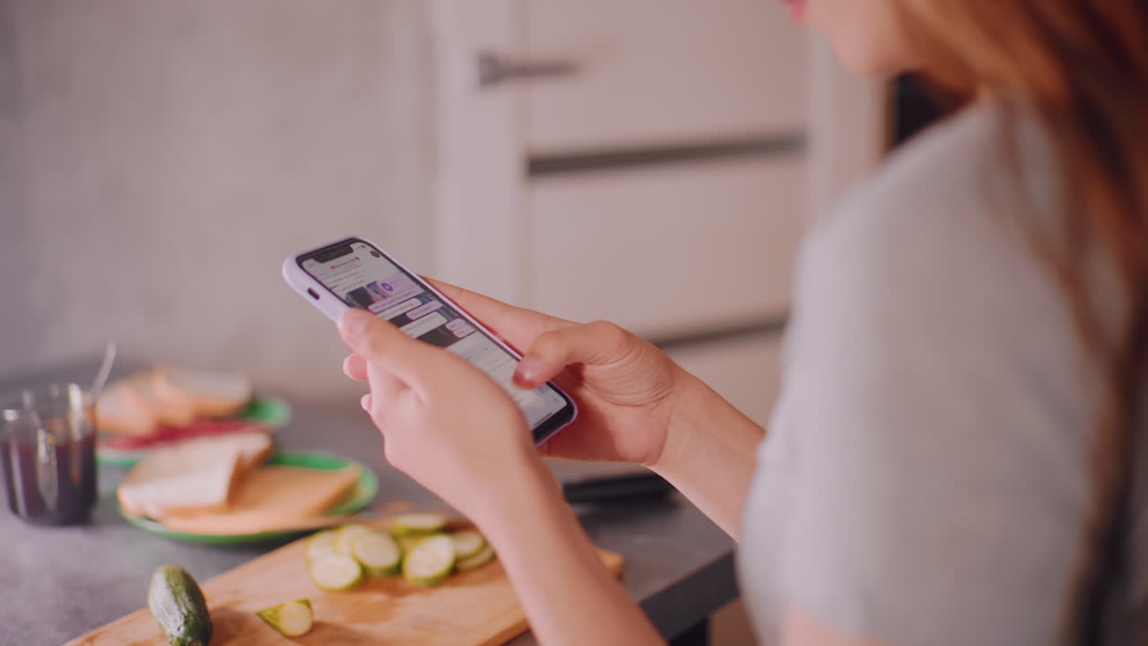 Close up of woman holding smartphone while reading chat messages in kitchen, sliced cucumber and bread on cutting board with coffee cup in background, blending digital lifestyle with daily routine