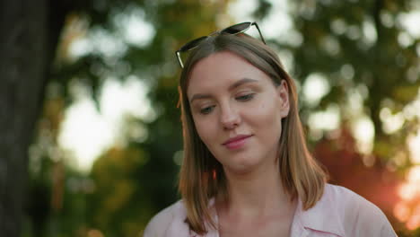 close-up of young woman looking up contemplatively with glasses resting on her head, focusing on something ahead, with background of trees and light filtering through them