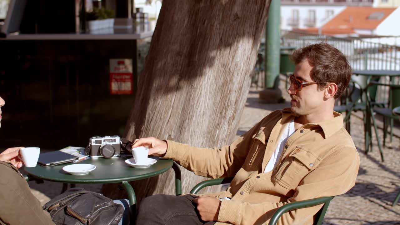 Candid moment in a sunlit European café square, as two people enjoy espresso and heartfelt conversation. Warm lighting casts soft shadows in a picturesque, tranquil setting