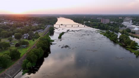 golden sunset at horizon between trenton, new jersey and morrisville,pa . reflection on delaware river with green island and bridge. aerial backwards wide shot. peaceful serene atmosphere in summer