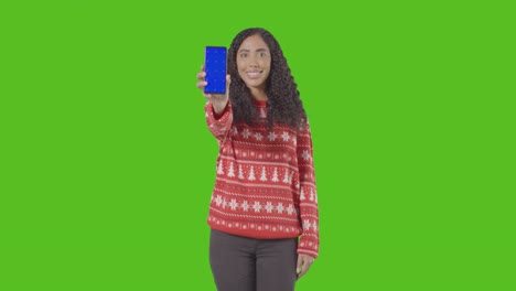 studio portrait of woman wearing christmas jumper holding up mobile phone against green screen