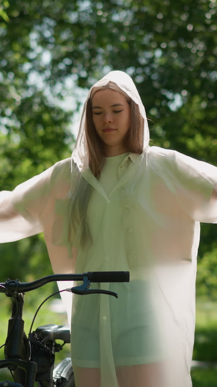 Young woman in translucent raincoat stretches her arms joyfully near bicycle, swaying her hands in sunny park surrounded by lush greenery