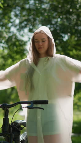 joven en impermeable translúcido extiende sus brazos alegremente cerca de la bicicleta, balanceando sus manos en un parque soleado rodeado de exuberante vegetación