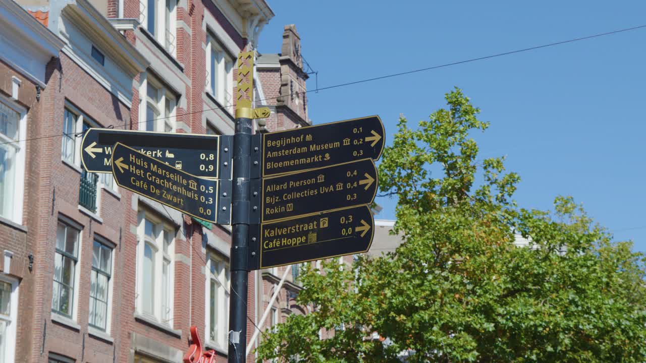 Black street signs near brick buildings and green trees under clear blue sky, static shot