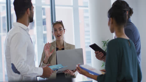 Mujer-De-Negocios-Caucásica-Sosteniendo-Una-Computadora-Portátil-Hablando-Con-Diversos-Colegas-De-Pie-En-Una-Reunión-De-Oficina