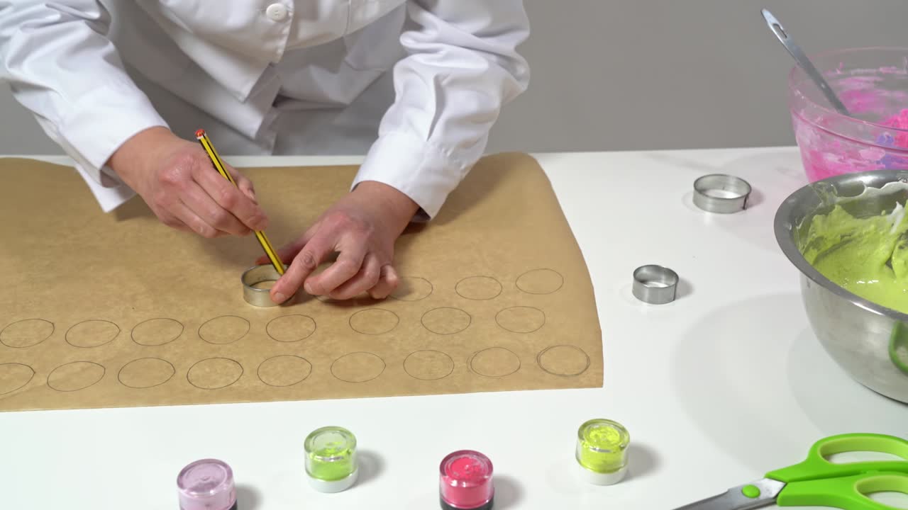 panadero que utiliza un lápiz y un pequeño cilindro de metal para hacer formas circulares en papel de pergamino, para ayudar en la fabricación de macarrones.