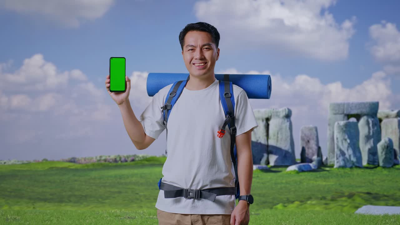 Asian Male Hiker With Mountaineering Backpack Smiling And Showing Mock Up Green Screen Smartphone While Traveling In Stonehenge