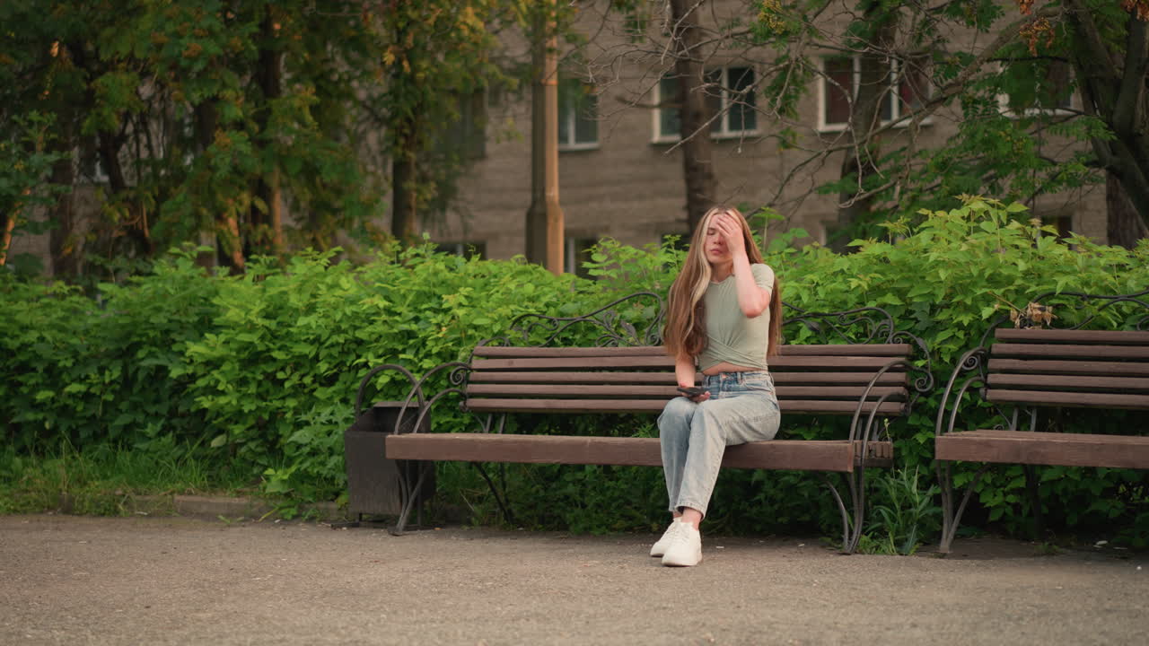 밖의 나무 벤치에 앉아있는 여인, 오른손으로 전화를 보고, 왼손으로 머리카락을 부드럽게 만지고, 배경에는 나무와 건물이 부분적으로 잎자루로 가려져 있습니다.