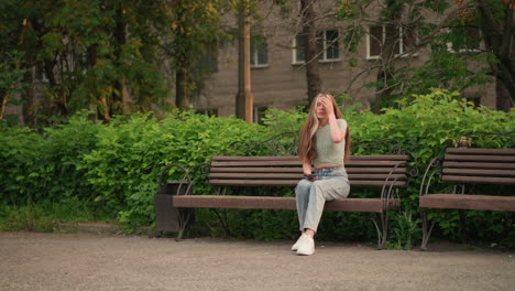 lady sitting on outdoor wooden bench, looking at phone in her right hand, gently touching her hair with her left hand, background includes, trees, and building partially obscured by foliage