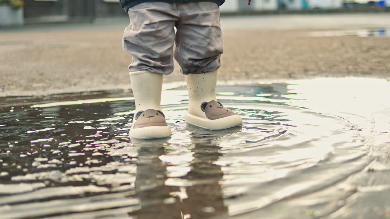 Close-up of children's feet in rubber boots. Cute two-year-old boy in a jacket and rubber boots walks on the street near puddles. Child in rubber boots runs through puddles