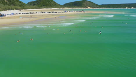 Aerial-4K-Drone-Over-Surfers-In-Turquoise-Ocean-Beside-Sandy-Beach-Shore-With-Cars-And-Camping,-Australia