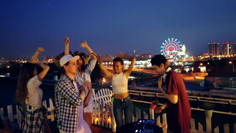 joyful dj is working with mixer at rooftop party enjoying music while young people girls and guys are dancing raising hands. beautiful view of night city is in background.