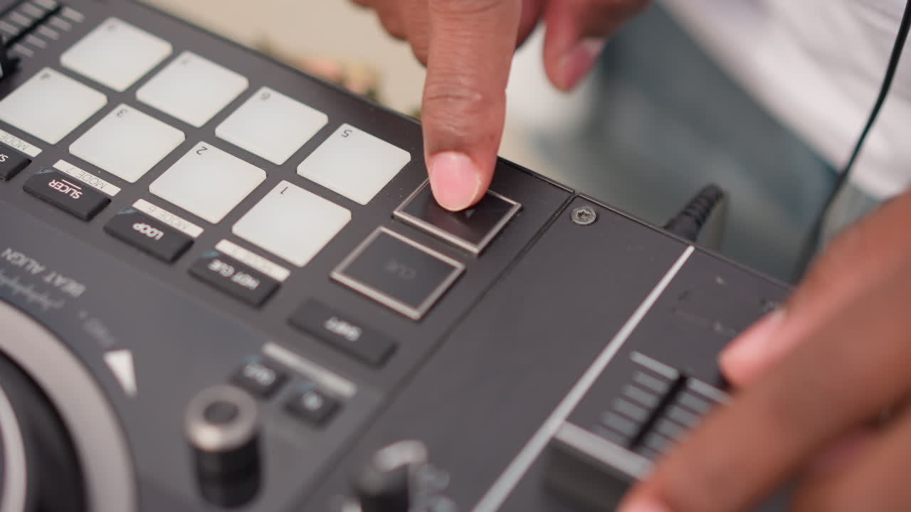 Gros plan des mains d'un DJ noir tapant sur les pads d'un contrôleur compact, cueillant des pistes, ajustant le fader et le bouton, éclairage intérieur chaleureux, création rythmique concentrée, ambiance de rue urbaine, roue jog et câble visibles