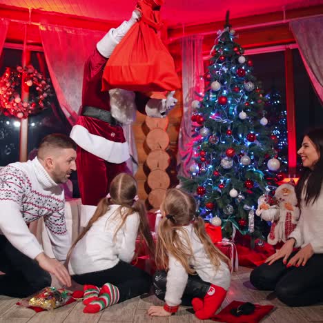 cheerful family sitting near a fireplace and meeting santa with a big bag. kids rejoicing when seeing presents. santa comes to the family and sits beside them under the christmas tree