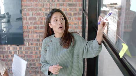 biracial casual businesswoman in discussion, brainstorming at glass wall in office, slow motion