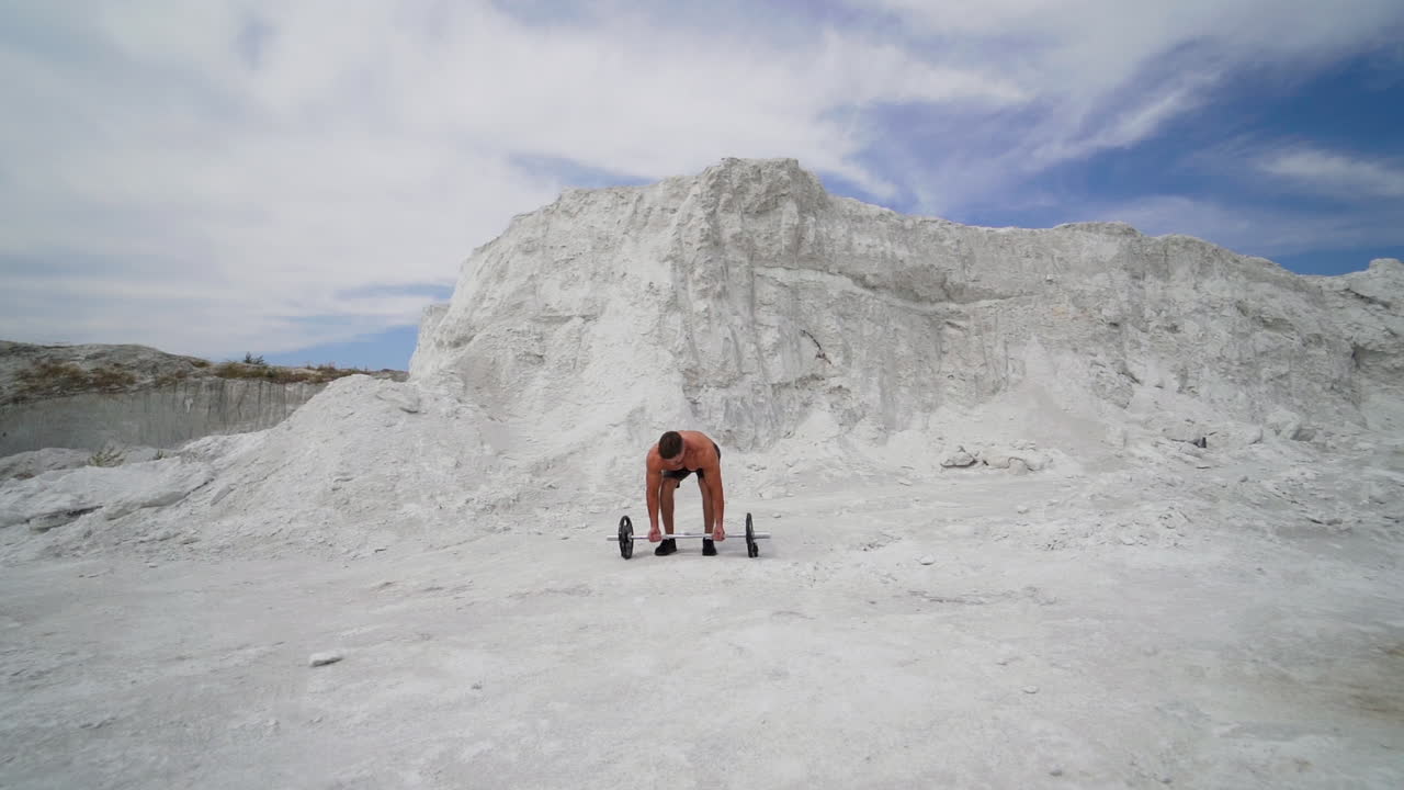 Strong shirtless athlete taking heavy barbell outdoors. Muscular man lifting barbell on white mountain background. Slow motion.