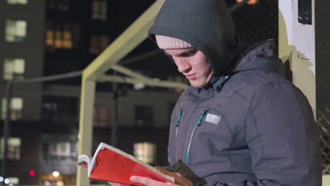 estudiante en chaqueta y gorra apoyado contra el poste de meta metálico centrado en la lectura de libros al aire libre por la noche bajo las luces de las calles de la ciudad urbana, momento pacífico de lectura en un entorno urbano