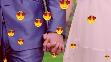 Multiple-heart-eyes-face-emoji-floating-against-mid-section-of-newly-married-couple-holding-hands