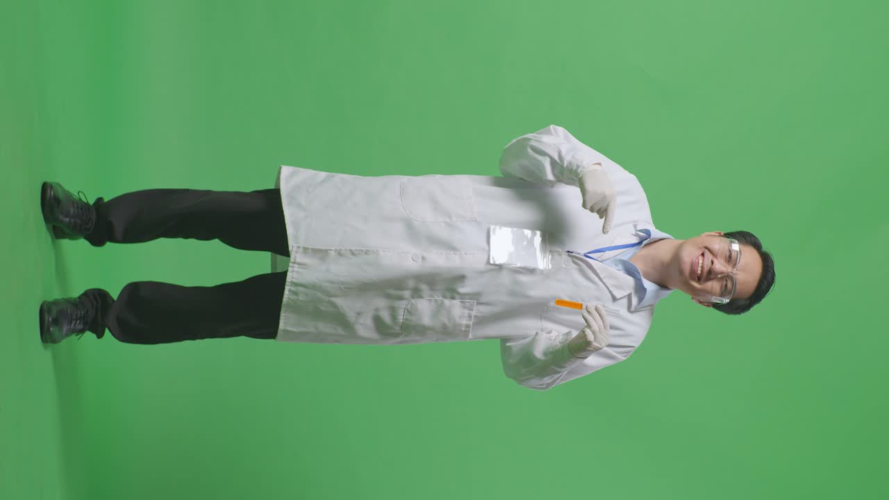Full Body Of Asian Man Scientist Smiling And Pointing To Orange Liquid In The Test Tube In His Hand While Standing On The Green Screen Background In The Laboratory