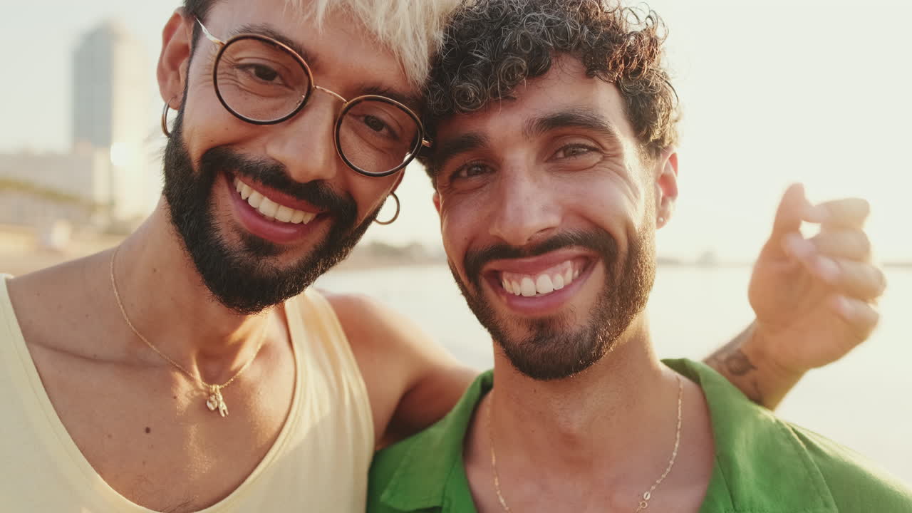 Coppia gay felice che si abbraccia e sorride alla fotocamera in riva al mare