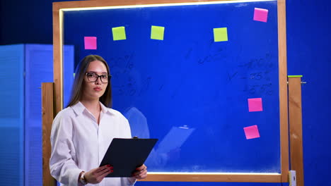 caucasian female in white shirt and glasses holds a clipboard. lady is getting ready for business presentation. glass board with stickers at backdrop.