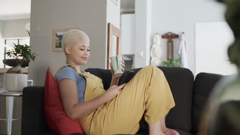 mujer biracial feliz usando un teléfono inteligente y sentada en el sofá con café en cámara lenta