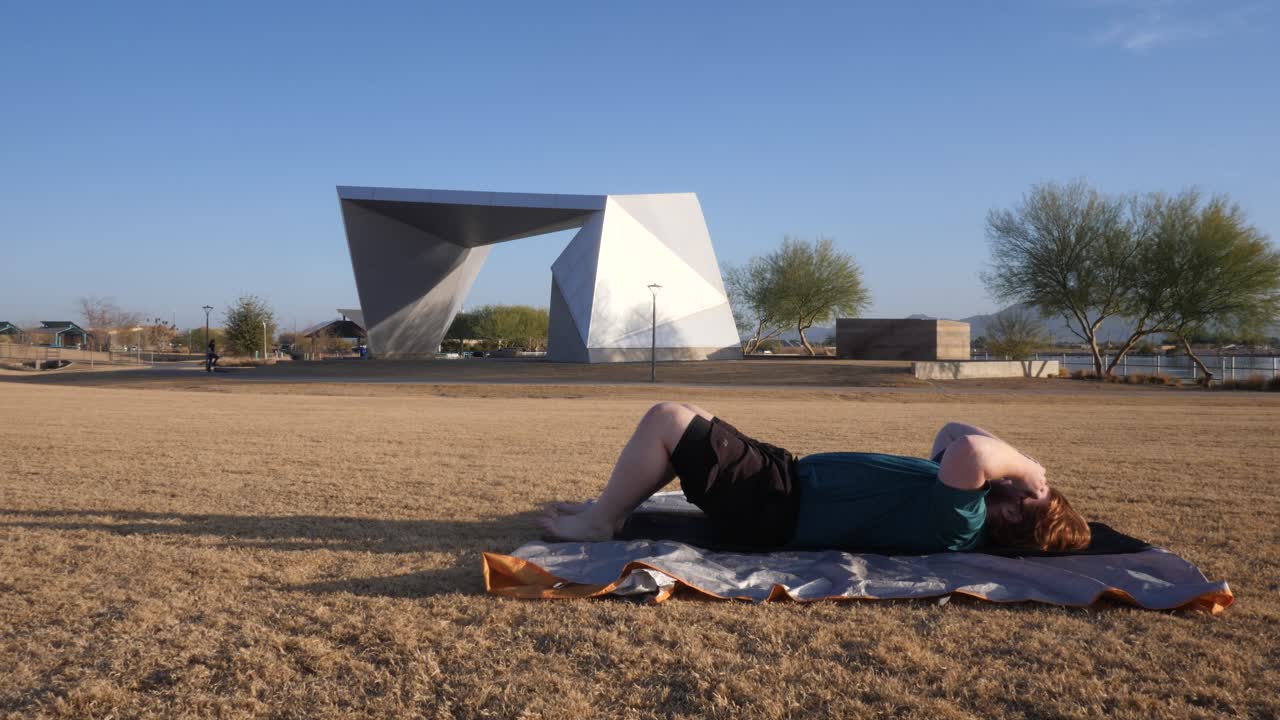 Overweight Red hair male doing sit ups in a park with an amphitheater behind them.
