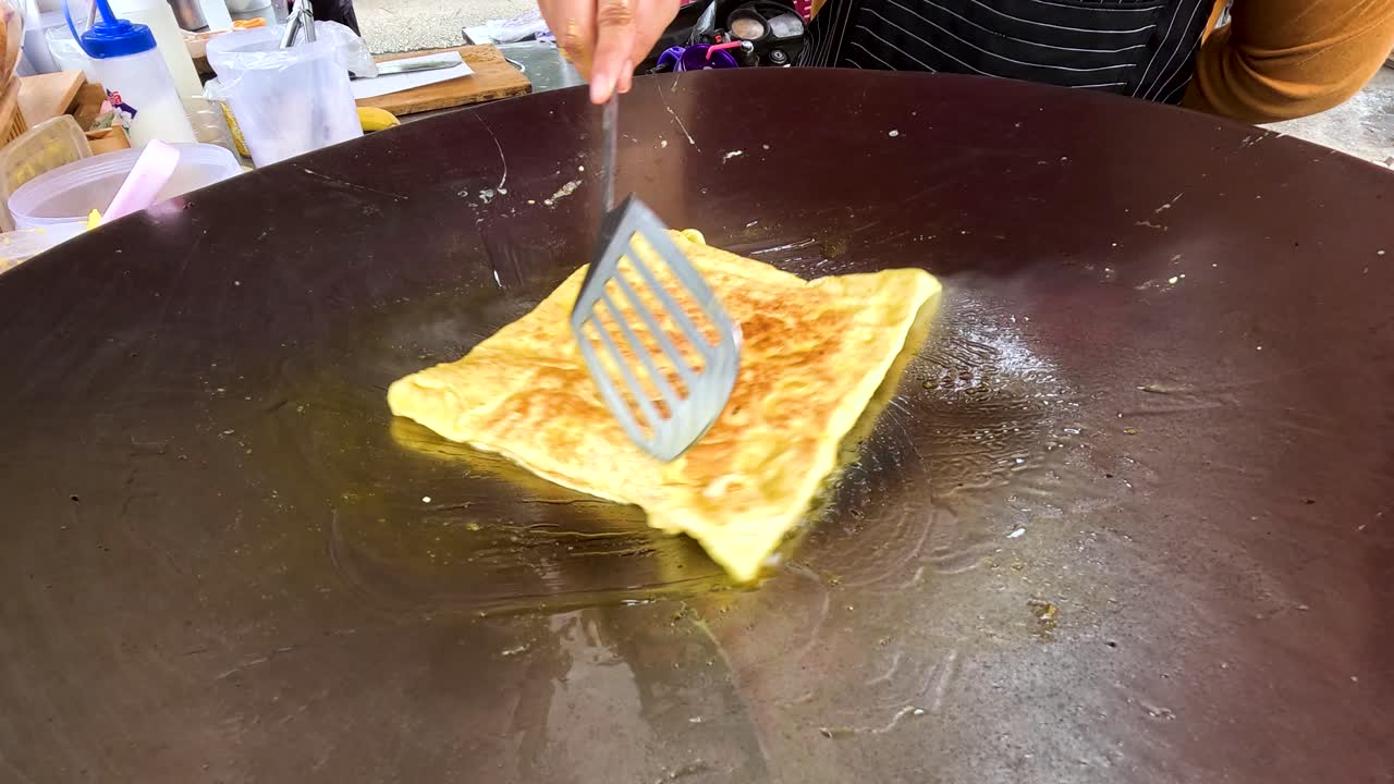 A street vendor expertly cooks fried roti on a hot griddle in Kanchanaburi, Thailand. The process highlights traditional techniques and vibrant street food culture