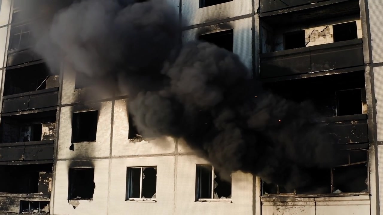 Fogo irrompendo na fachada de apartamento em andar intermediário, enviando fumaça preta através de janelas quebradas, brasas