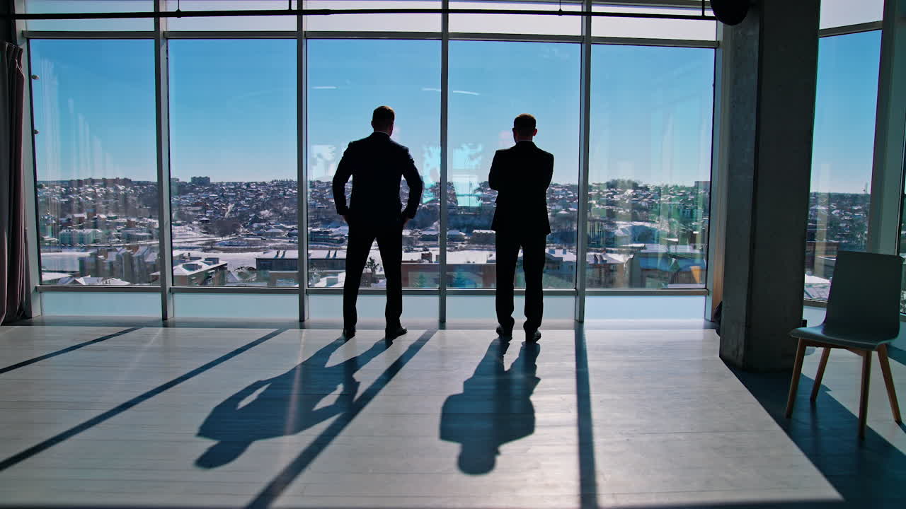 Businessmen in dark suits talking during break. Office workers standing near large windows and looking on city. Rear view.