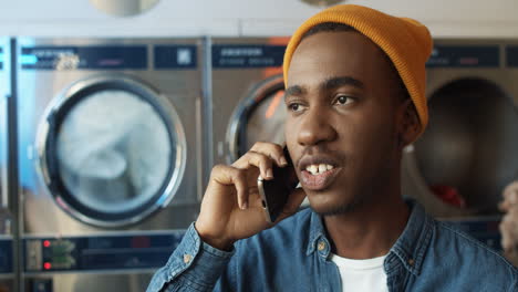 Close-Up-Of-Young-Cheerful-Handsome-Guy-In-Yellow-Hat-Talking-On-Mobile-Phone-And-Smiling-In-Laundry-Service-Room-Happy-Man-Speaking-On-Cellphone-In-Washhouse