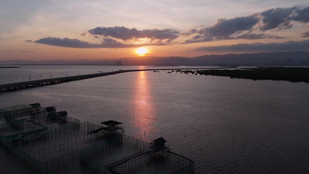 Drone view of floating aquaculture fish cages silhouetted against a golden sunset. Traditional bamboo fish pens situated near the modern Davao Coastal Road with sun reflection on the calm sea