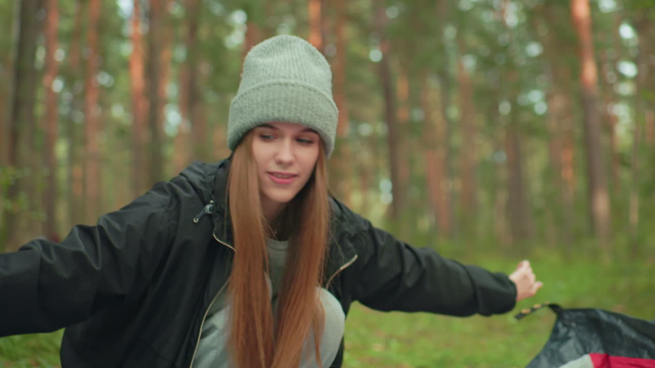 Close up of cheerful young lady wearing gray beanie and black hoodie smiling gently while bending forward during outdoor camping setup in forest, surrounded by tall pine trees and lush green scenery