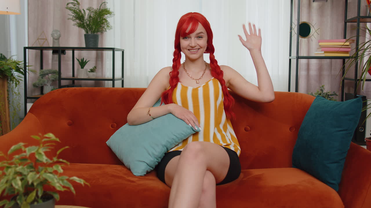 빨간 머리 소녀는 카메라에 친절하게 미소 짓고, 손을 흔들며, 안 ⁇ 하세요, 안 ⁇ 하세요, 소파에서 집에서 인사합니다.