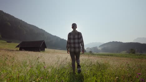 A-woman-walks-through-a-sunny-field-with-mountains-and-a-rustic-cabin-in-the-background