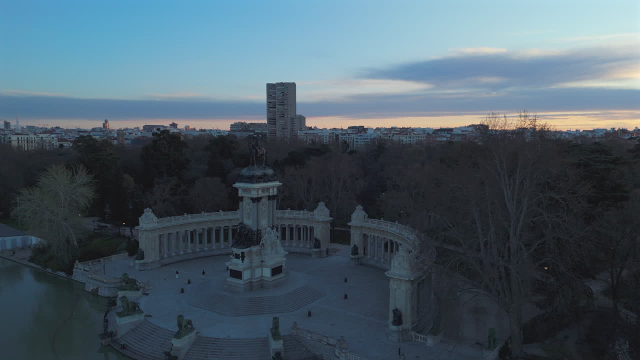 Drone footage circling the Alfonso XII monument in Retiro Park at sunrise, showcasing Madrid’s skyline, city buildings, and a vibrant morning sky with warm sunlight reflecting over the horizon