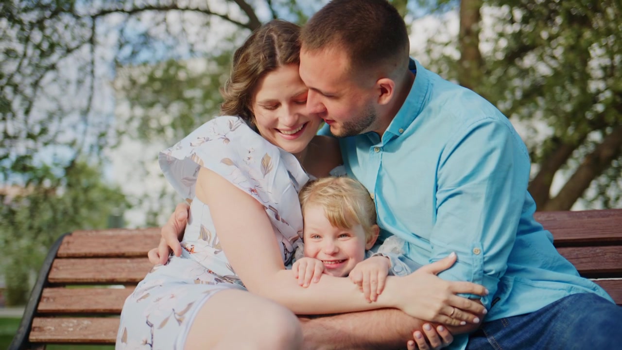 Una famiglia felice di tre persone siede su una panchina di legno sotto alberi verdi. La madre sorride calorosamente mentre tiene in braccio il bambino. Il padre guarda teneramente il bambino. Il bambino guarda la telecamera con innocente curiosità.