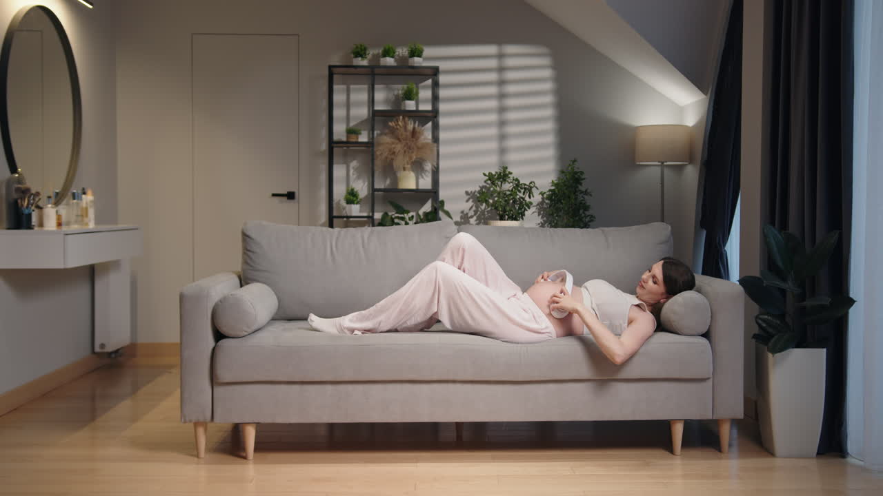Pregnant woman lying on sofa wearing headphones on belly listening music enjoying pregnancy moments