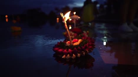 loy krathong festival: colorful krathong leaning left, floating on the dark blue water on the lake with a burning candle and instance on top