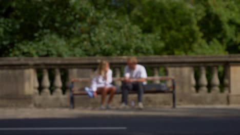 Defocused-Shot-of-People-and-Traffic-Moving-In-Front-of-Couple-Sat-On-Bench-01